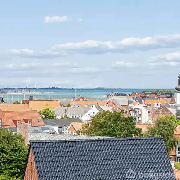 Byens tage dominerer forgrunden, mens en kirke med et spir står ud blandt hustagene. I baggrunden ses en rolig blå havnefront under en skyet himmel.