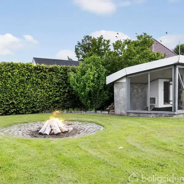 Et bål brænder på en rund bålplads omgivet af græs. Ved siden af er en lille moderne pavillon med glasdøre, alt placeret i en indhegnet have med høje hække.