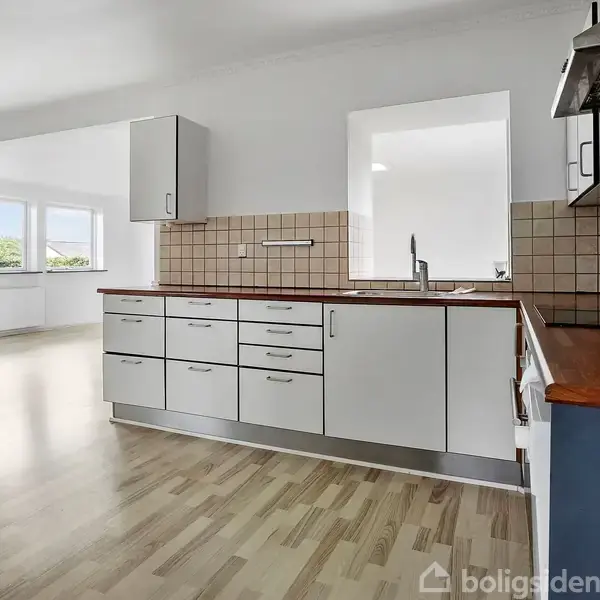 Et moderne køkken med hvide skabe og en stålgrå ovn, står i et lyst rum med trægulv. Store vinduer i baggrunden tillader naturligt lysindfald.