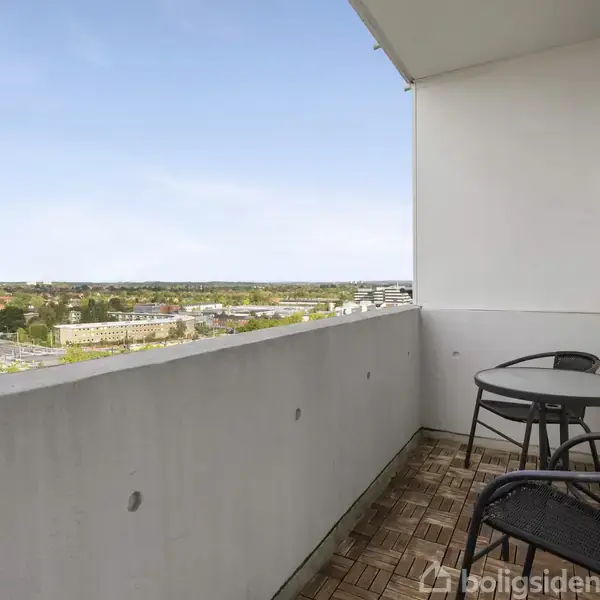 En balkon med to sorte kurvestole og et lille rundt bord. Udsigt over byområde med træer og bygninger under en klar blå himmel.