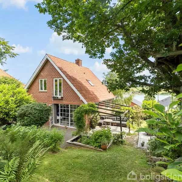Rødt murstenshus står i en frodig have med græsplæne og træer. En glasveranda er ved siden af huset, og en stor træterrasse strækker sig ud fra bygningen.