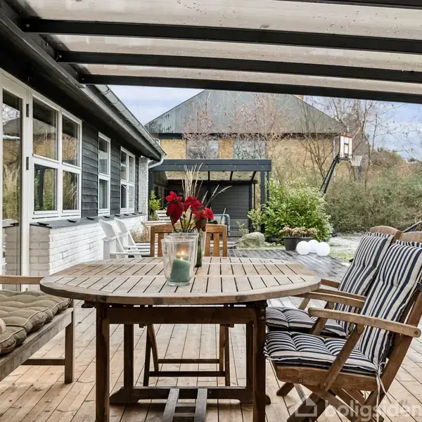 Rundt træbord med tændt lys i glas, omgivet af stole og bænk, står på en overdækket terrasse ved et hus. Haven i baggrunden er delvist anlagt med planter og buske.