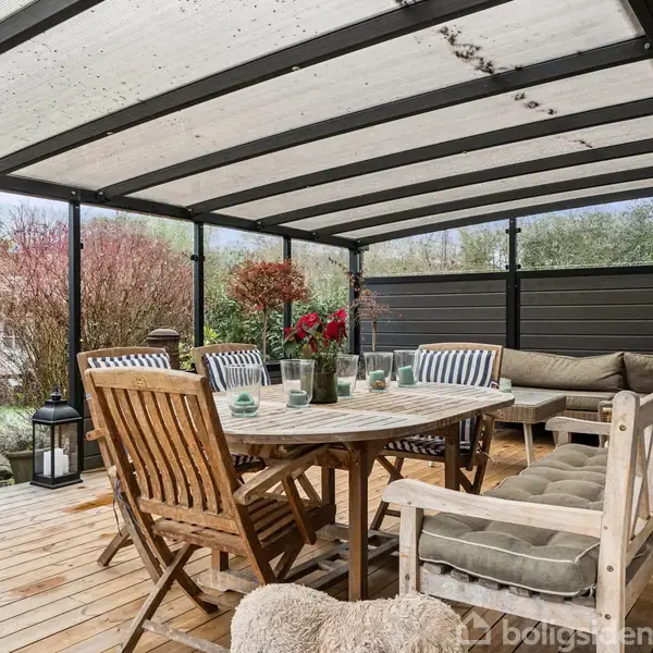 Et overdækket terrasseområde med træbord og stole; bordet er pyntet med glas og stearinlys. I baggrunden ses en sofa og planter, omgivet af en have med buske.