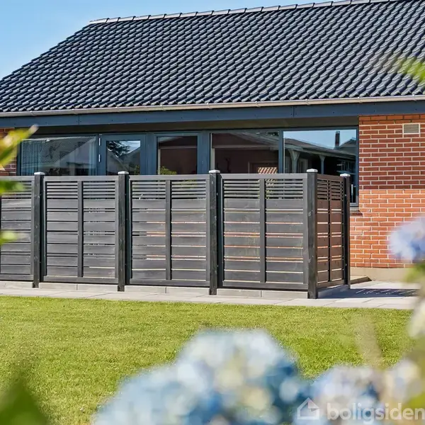 Et rødt murstenshus med mørkt tag ses i baggrunden. Foran huset er der et sort, moderne plankeværk. Grøn græsplæne og blomster fylder forgrunden.