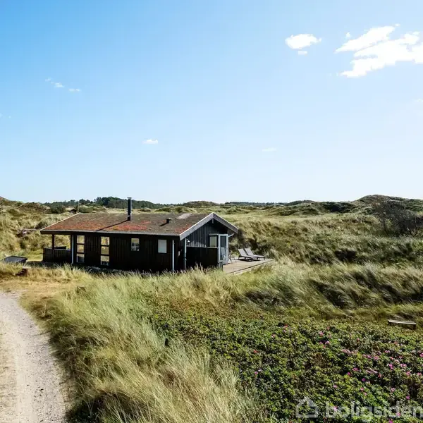 Et lille, sort træhus ligger stille mellem grønne klitter under en blå himmel med få skyer. En smal grusvej snor sig forbi huset gennem det naturlige landskab.