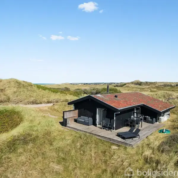 Et sort sommerhus står stille på en træterrasse omgivet af grønne klitter under en klar blå himmel.