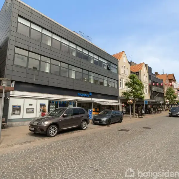 Bygning med store vinduer står langs en brostensbelagt gade. Foran holder flere biler parkeret. Tekst på bygningen: "Nordea". Omgivet af andre bygninger og små butikker på en solrig dag.