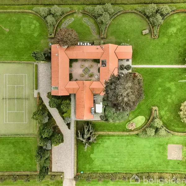Stort hus med rødt tag set oppefra. Huset omgiver en central gårdhave. Til venstre ses en tennisbane, omgivet af velholdte grønne plæner og træer.