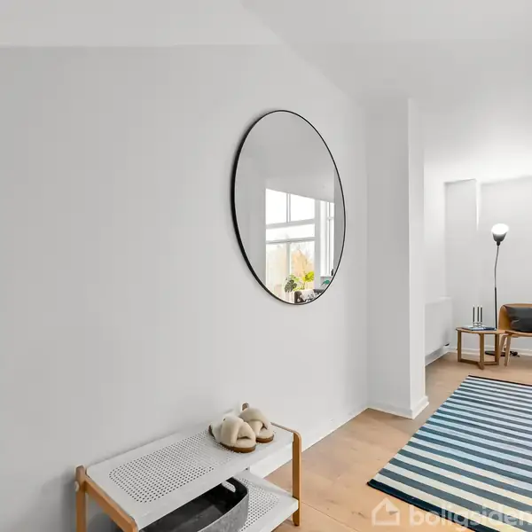 Rundt spejl hænger på væg; reflekterer lys. Under spejlet, lille hylde med hjemmesko. I baggrunden, stue med gulvlampe, stol, plante og flerfarvet tæppe på trægulv.