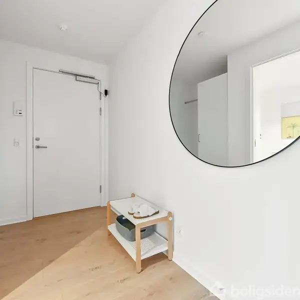 Stor, rund vægspejl hænger i en lys entré. En lille hvid hylde med to niveauer står på parketgulvet ved siden af en hvid dør.