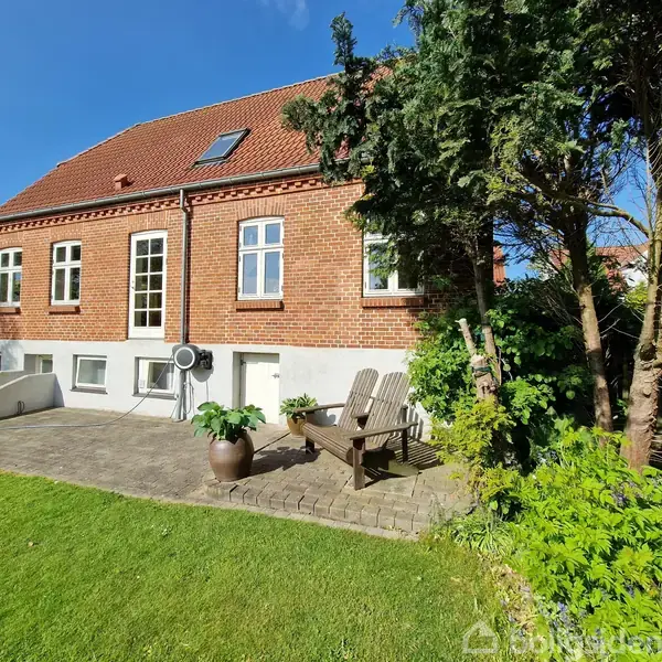 Rødt murstenshus står stille i en have med fliser og græsplæne. En bænk og planter omkranser området. Klar blå himmel danner baggrunden.