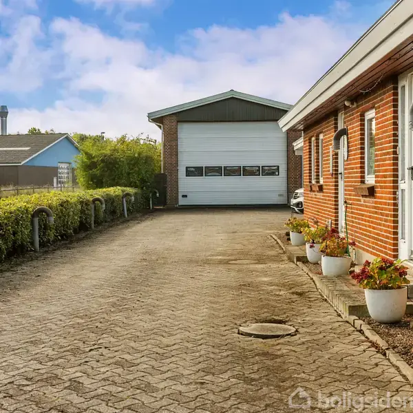En lang indkørsel fører op til en garage med hvid port, omgivet af et rødstenshus med hvide døre. Blomsterkrukker står ved siden af huset, og en hæk løber parallelt.