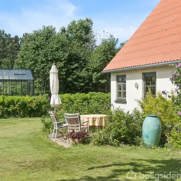 Hus med rødt tag står ved grøn have med borde og stole. Parasol er ved siden af terrassen. Drivhus kan ses i baggrunden omgivet af buske og træer.