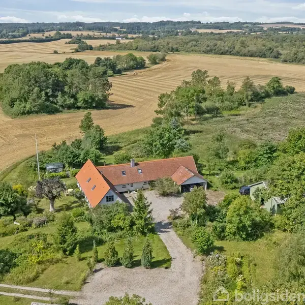 Et hus med rødt tag ligger omgivet af frodig natur og marker. Stier og omgivende træer skaber en landlig atmosfære, mens kuperet terræn ses i baggrunden.