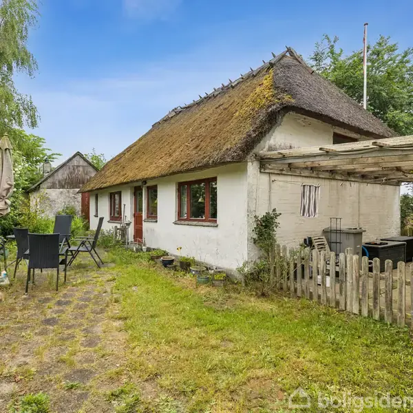 Hus med stråtag står i en grøn have. Et udendørs spisebord og stole står placeret på en grusstien. Omkring huset er der træer og en terrasse med pergola.