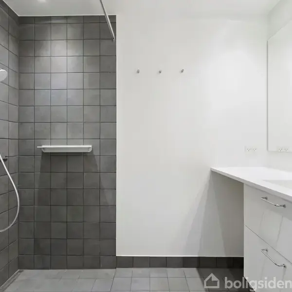 Brusekabine med bruser i grå fliser. Ved siden af er der en hvid vask med skab og spejl. Rummet har lyse vægge og et enkelt udtryk.