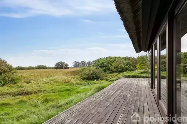 En træterrasse grænser op til et hus med stråtag, hvor den åbner sig mod en åben udsigt over en græsmark med spredte buske og træer under en klar blå himmel.