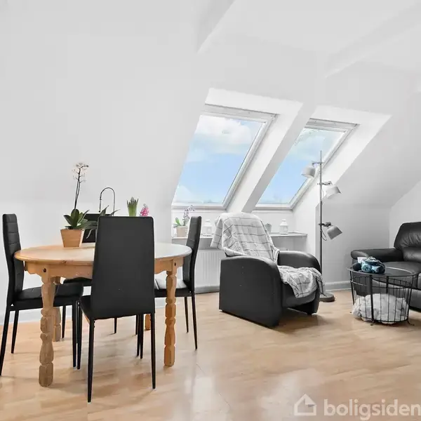 Et lyst stueområde med skråvinduer viser en spiseplads med rundt træbord og sorte stole. Ved siden af står en lænestol og sort sofa, omgivet af lyse trægulve.