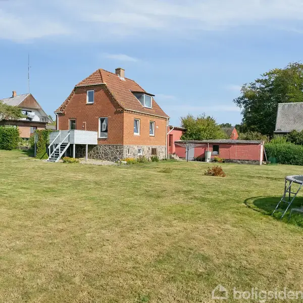 Et rødt murstenshus med en lille trappe står i en græsplæne med buske og træer i baggrunden. En trampolin er placeret i højre del af billedet.