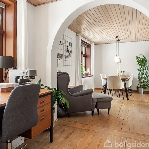 Kontorbord med computer og lampe, stående mod et vindue, i et rum med bueåbning til hyggelig spisestue med spisebord, stole og planter. Miljøet er lyst og moderne.