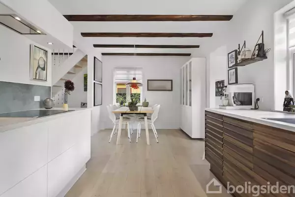 Køkken-alrum med hvide skabe og træbjælker i loftet. Et spisebord med stole står centralt, omgivet af kunst og planter. Store vinduer lyser rummet op.