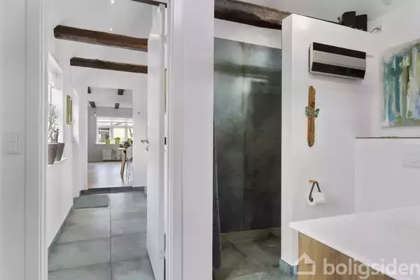 Badeværelse med sort brusekabine og hvidmalede vægge. Til højre ses et toiletpapirholder og en moderne radiator. I baggrunden fører en gang til en spisestue med vinduer.