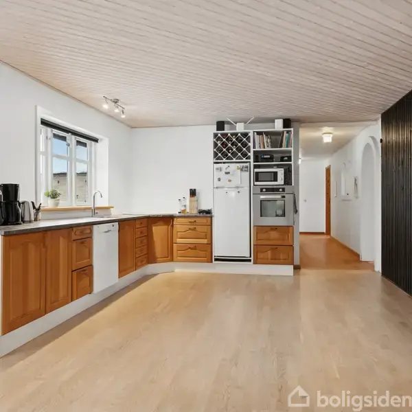 Køkken med træskabe og sort bordplade, hvorpå en kaffemaskine står. Indbygget ovn og mikroovn placeret ved siden af køleskab. Store vinduer åbner op til naturligt lys.