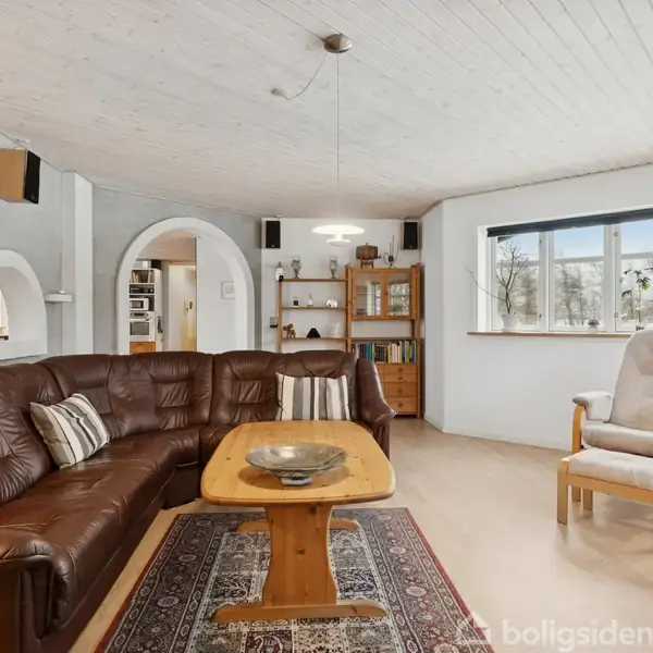 En stue med en brun hjørnesofa og et træsofabord står på et orientalsk tæppe. To lænestole er placeret ved et stort vindue. En reol står nær en hvælving.