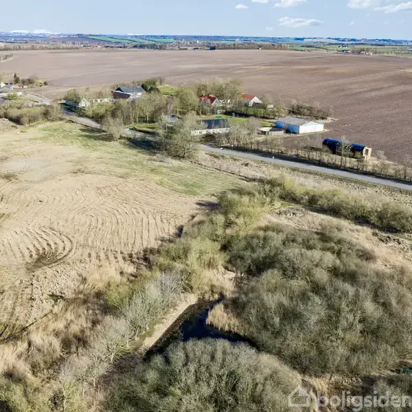 Landbrugsjord strækker sig over et stort, åbent område, med spor af traktorfurer. Spredte bygninger og siloer er synlige i baggrunden. Skyfri himmel over markerne.