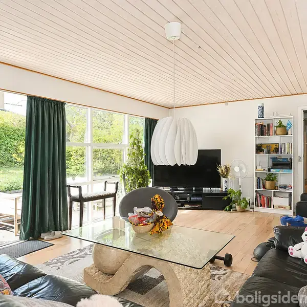 Stue med glasbord omgivet af sorte lædersofaer. En stor lampe hænger fra træloftet. Bøger og dekorationer er på hylderne. Store vinduer åbner mod en grøn have.