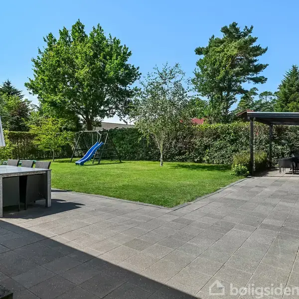 En have med en flisebelagt terrasse, borde og stole under en parasol. En blå rutsjebane står på græsplænen omgivet af træer og buske, med et halvtag i baggrunden.