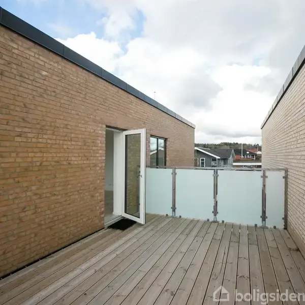 Dør står åben på en tagterrasse med trædæk, omgivet af røde murstensvægge. Gennemsigtig glasrækværk afskærmer kanten. Himlen er overskyet, og bygningsdele kan ses i baggrunden.
