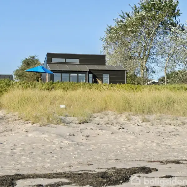 Et moderne sommerhus med store vinduer og en blå parasol står blandt sandklitter og græs nær stranden, omgivet af træer og blå himmel.