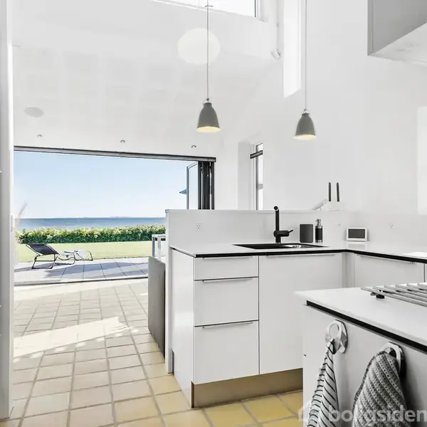 Et moderne køkken med hvide skabe og sorte bordplader leder ud til en terrasse med udsigt over havet. To liggestole står ved den åbne terrassedør, omgivet af grønne buske.