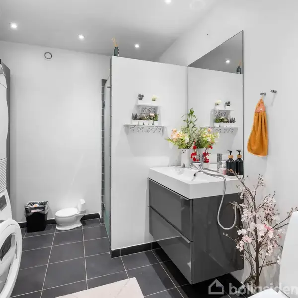 Vaskemaskine og tørretumbler er stablet i et badeværelse med mørke flisegulve. En håndvask med blomsterdekorationer står ved siden af, mens håndklæder hænger på væggen.