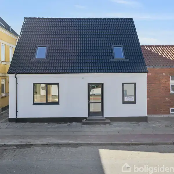 En hvid murstenshus med sorte tagsten og to ovenlysvinduer ses i en bymæssig gade omgivet af andre huse. Et skilt i vinduet siger "Til salg".