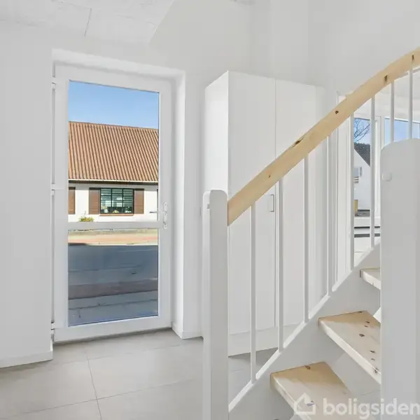 En lyse trappeopgang med lyst trægulv fører opad. En glasdør åbner til gaden, hvor et hus med rødt tag ses. Rummet er hvidt og minimalistisk indrettet.