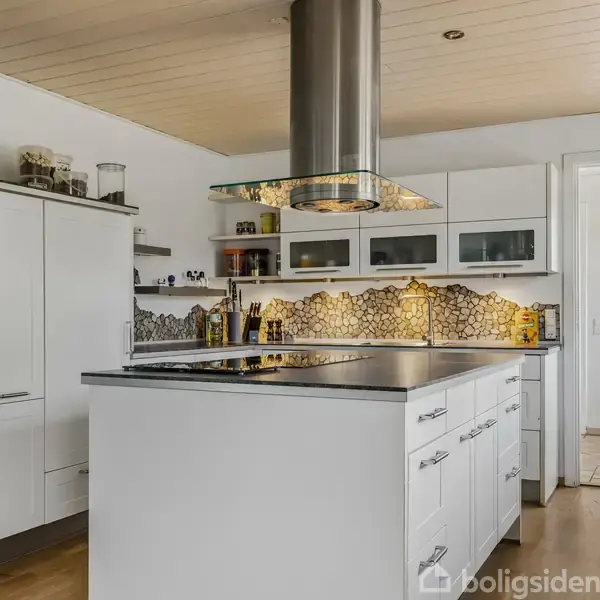 Et moderne køkken har en hvid køkkenø med integreret kogeplade og emhætte. Omkringstående skuffer og skabe er i hvidt design. Væggene har mosaikfliser, og der er trægulv.