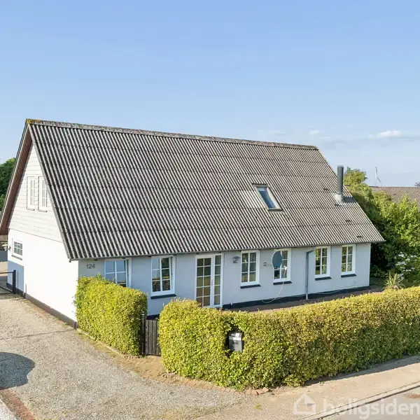 Et hvidt hus med gråt tag står omgivet af grønne hække i en forstadslignende omgivelser. Ved siden af huset holder en sort bil på en grusindkørsel.