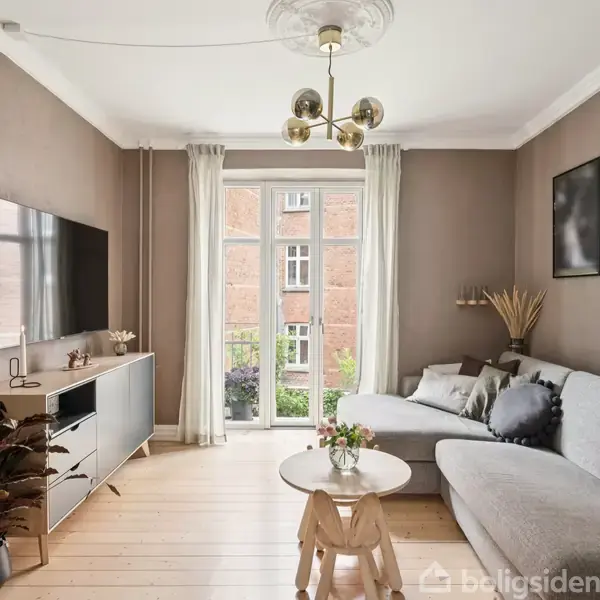 En lys stue med en grå hjørnesofa ved en brun væg, dekoreret med billedrammer. En rund træbord med blomster i en vase står foran sofaen. Store vinduer til venstre.