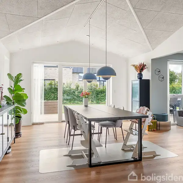 Spisebord med stole er midtpunktet, omgivet af planter, kunst på væggene, og store glasdøre der åbner ud til en terrasse. Loftslamper hænger over bordet, skaber en lys og indbydende atmosfære.