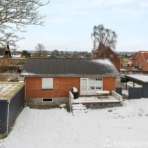 Et rødt murstenshus står i et snedækket boligområde med flere huse i baggrunden. En terrasse foran huset har et havebord og -stol. Træer uden blade omgiver området.