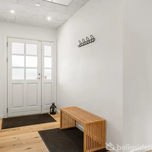 En træbænk står i en entré med hvide vægge. En dør med glasruder ses i baggrunden, omgivet af måtter og en simpel knagerække på væggen.