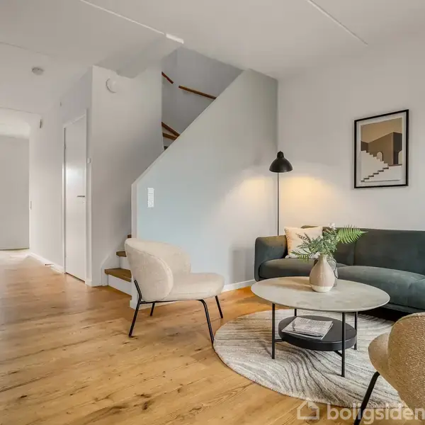 En moderne stue med en grå sofa og to beige stole står omkring et rundt sofabord. En trappe fører opad ved siden af, og en spisestue kan ses i baggrunden.