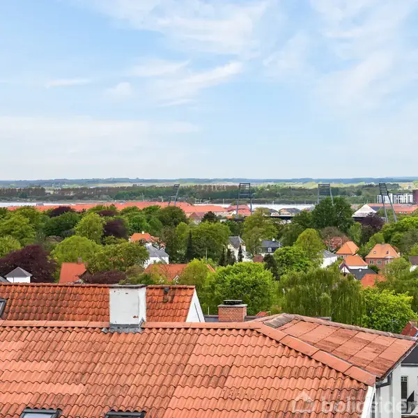 Rødt tag hviler stille over rækkehuse omgivet af frodige træer og grønne parker, med en stor vandmasse og bysilhuet i baggrunden under en klar, blå himmel.