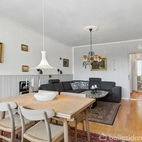 Stue med gråt hjørnesofaarrangement og sofabord på tæppe. Træspisebord med fire stole under en hvid lampe. Væggene er pyntet med malerier; en bogreol står i hjørnet.