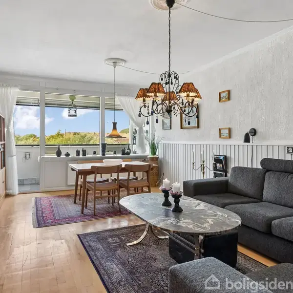 En stue med en stor grå sofa står mod en hvid væg. Foran er et ovalt sofabord. Til venstre ses en bogreol, et spisebord ved et vindue med udsigt til naturen.