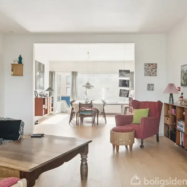 Stue med et sofabord og sort sofa i forgrunden, lys træhylde med bøger til højre. I baggrunden ses et spisebord med stole i et lyst rum.