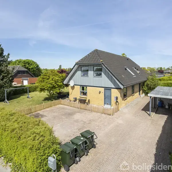 Et tofamiliershus med blå tag og gul facade står stille. Det er omgivet af en have og en stor indkørsel med carport. Skraldespande står ved hegnet.