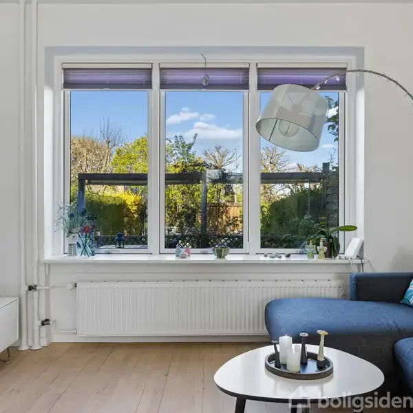 Sofa med farverige puder står ved et vindue med udsigt til en have. Et rundt bord har dekorative genstande, og en gulvlampe hænger over den.